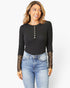 A woman modeling a black long-sleeve ribbed top with decorative gold buttons down the front and sheer lace cuffs, paired with blue jeans, against a plain white background.