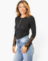 A woman modeling a black long-sleeve ribbed henley top with gold buttons and sheer black lace sleeves, paired with light blue jeans, against a plain white background.