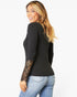 A woman wearing a black long-sleeve ribbed top with intricate black lace detailing on the lower sleeves, paired with light blue jeans, photographed against a solid white background.