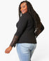 A woman modeling a black long-sleeve ribbed knit top with sheer black lace sleeves, paired with light blue jeans, against a plain white studio background.