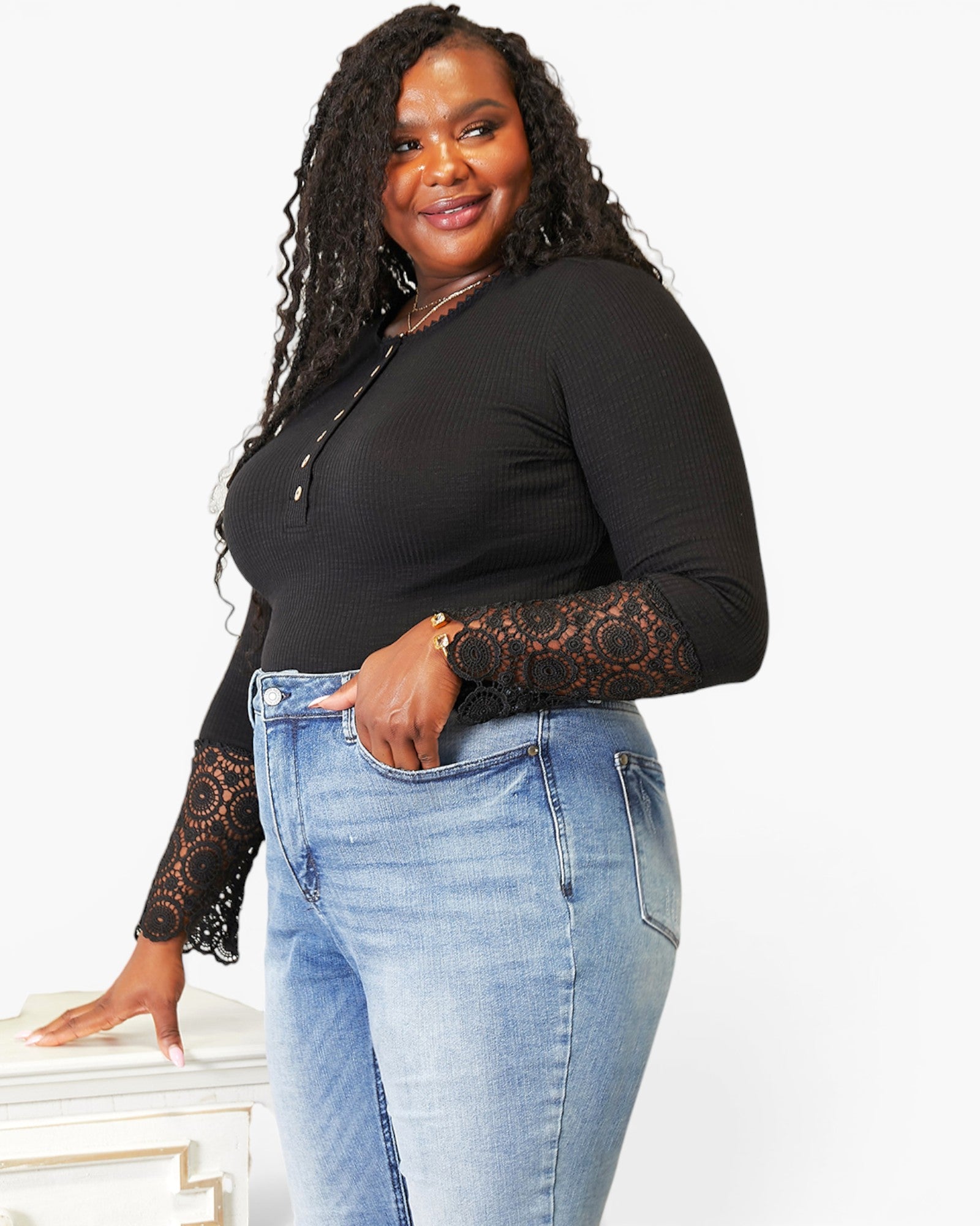 A woman modeling a black long-sleeve top with black lace sleeves and light blue jeans against a plain white background.