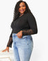 A woman modeling a black long-sleeve top with black lace sleeves and light blue jeans against a plain white background.