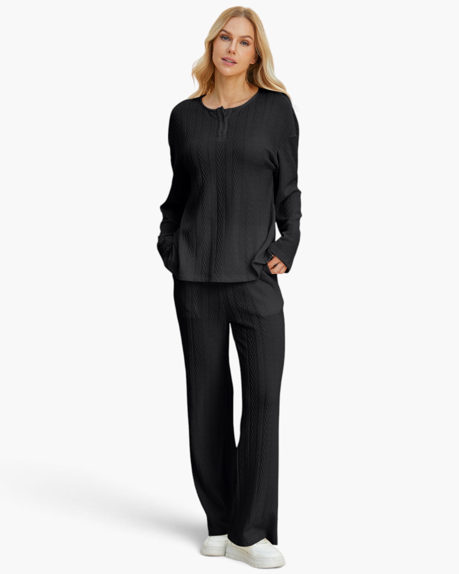 A woman modeling a matching black textured knit lounge set consisting of a long-sleeve top and wide-leg pants, paired with white sneakers against a light gray studio background.