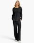 A woman modeling a matching black textured knit lounge set consisting of a long-sleeve top and wide-leg pants, paired with white sneakers against a light gray studio background.