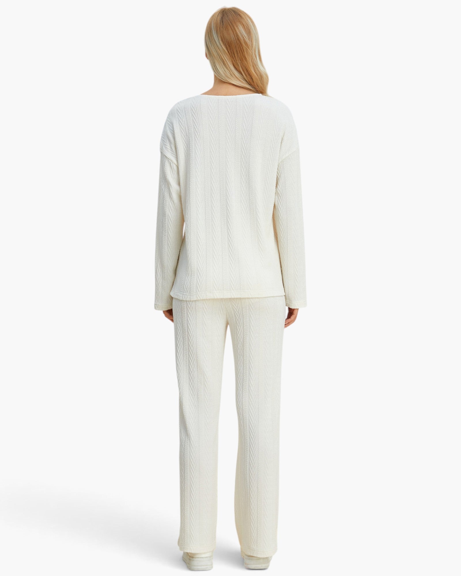 A woman viewed from the back wearing a cream-colored ribbed knit lounge set consisting of a long-sleeve top and matching wide-leg pants, set against a plain white studio background.
