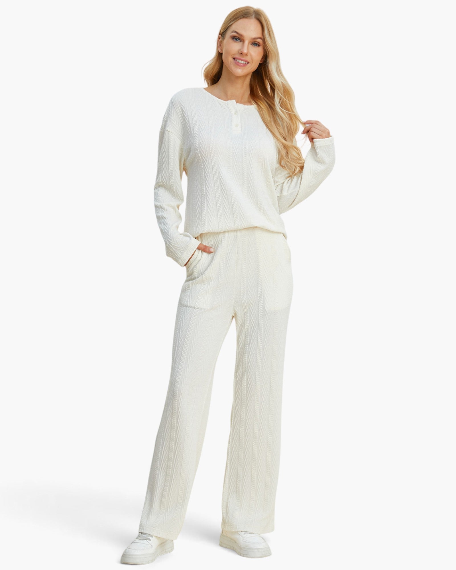 A woman modeling a matching cream-colored knit lounge set consisting of a long-sleeve henley top and wide-leg trousers, paired with white sneakers against a plain white studio background.