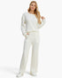 A woman modeling a matching cream-colored knit lounge set consisting of a long-sleeve henley top and wide-leg trousers, paired with white sneakers against a plain white studio background.
