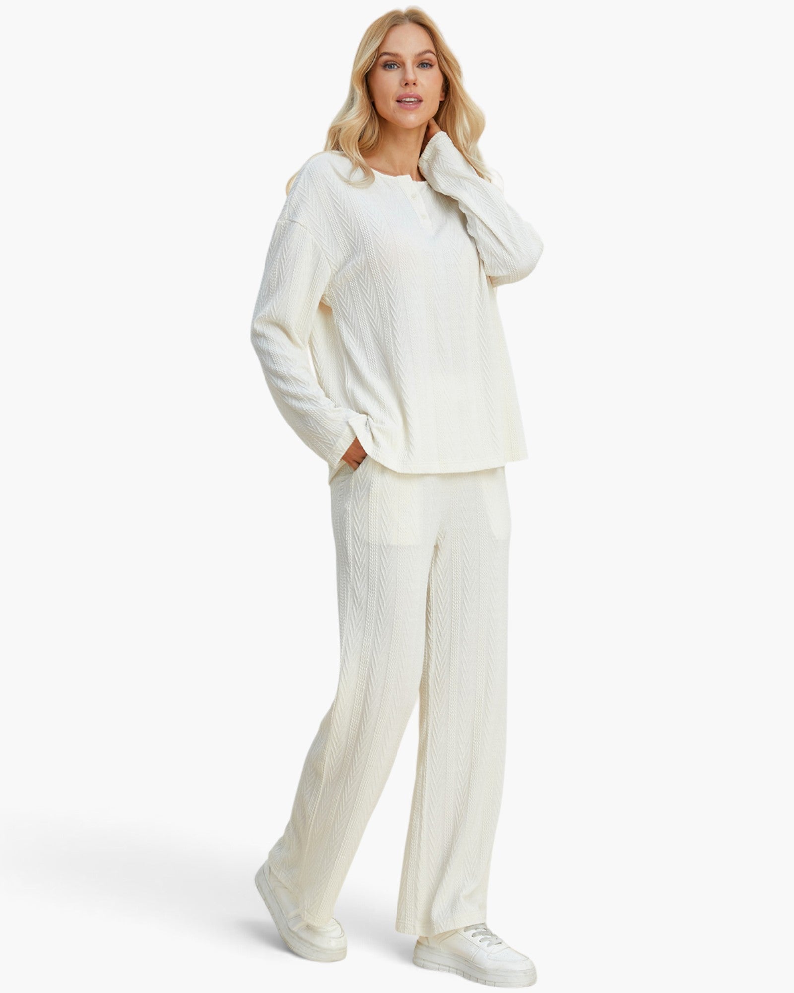 A woman modeling a matching cream-colored ribbed knit lounge set, featuring a long-sleeve top and wide-leg trousers, paired with white sneakers against a light gray studio background.