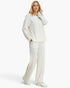 A woman modeling a matching cream-colored ribbed knit lounge set, featuring a long-sleeve top and wide-leg trousers, paired with white sneakers against a light gray studio background.