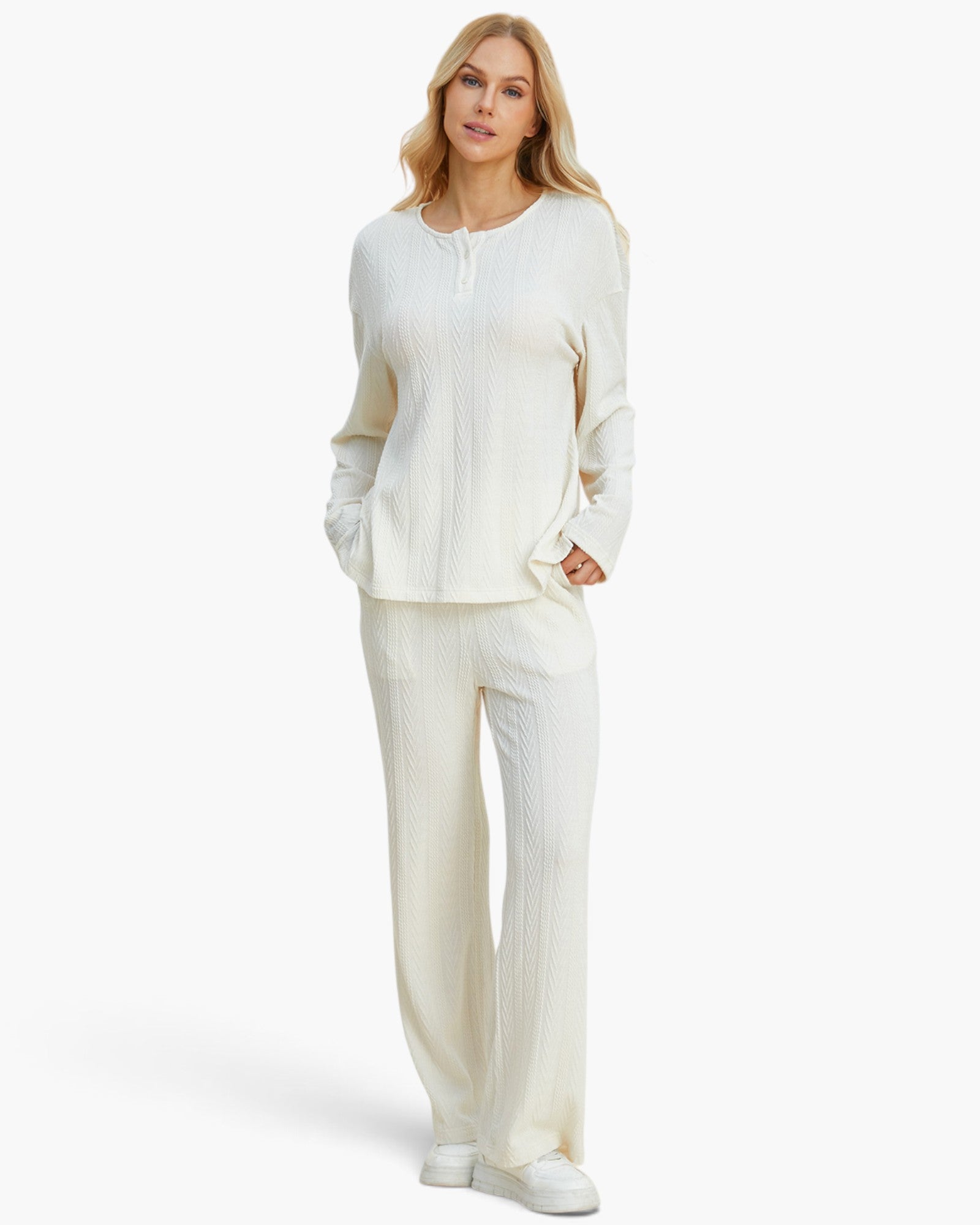 A woman modeling a matching cream-colored knit lounge set consisting of a long-sleeve henley top and wide-leg trousers, paired with white sneakers against a plain white studio background.