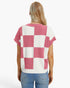 A woman viewed from the back wearing a pink and white checkered short-sleeve knit top with blue jeans, against a plain white studio background.