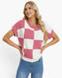 A woman modeling a short-sleeve knit top with a pink and off-white checkerboard pattern, paired with blue jeans, against a plain white studio background.