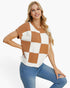 A woman modeling a short-sleeve knit top with a brown and off-white checkerboard pattern, paired with blue jeans, against a plain white background.