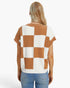 A woman viewed from the back wearing a short-sleeve knit top with a large checkered pattern in warm tan and off-white, paired with light blue jeans against a plain white studio background.