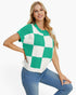 A woman modeling a short-sleeve knit top with a green and white checkerboard pattern, paired with blue jeans, against a plain white background.