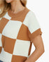 A close-up of a woman wearing a short-sleeve knit sweater with a large check pattern in warm camel brown and off-white. The sweater features a ribbed crew neck and sleeve cuffs, shown against a plain white background.