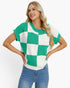 A woman modeling a short-sleeve knit top with a green and white checkerboard pattern, paired with blue jeans, against a plain white studio background.
