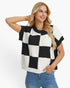 A woman modeling a short-sleeve knit top with a bold black and white checkerboard pattern, paired with blue jeans, against a plain white studio background.