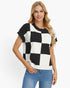 A woman modeling a short-sleeve knit top with a bold black and white checkerboard pattern, paired with blue jeans, against a plain white studio background.