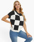 A woman modeling a short-sleeve knit top with a bold black and white checkerboard pattern, paired with blue jeans, against a plain white studio background.