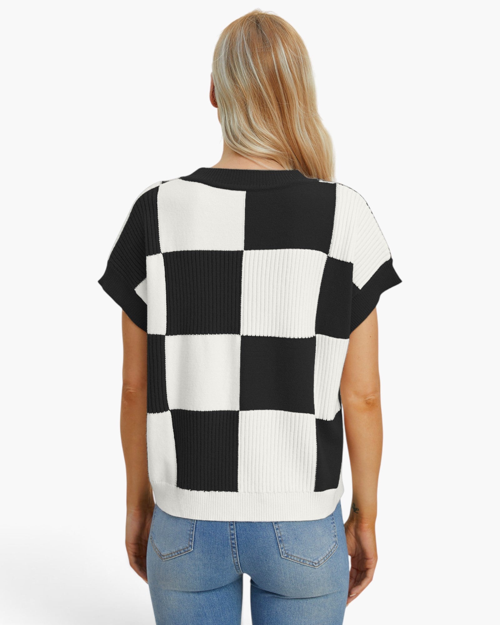 A woman viewed from the back wearing a short-sleeve knit top with a bold black and white checkerboard pattern, paired with light blue jeans, against a plain white studio background.