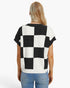 A woman viewed from the back wearing a short-sleeve knit top with a bold black and white checkerboard pattern, paired with light blue jeans, against a plain white studio background.