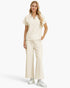 A woman modeling a matching cream-colored two-piece set consisting of a short-sleeve collared top and wide-leg drawstring pants, paired with white sneakers against a light gray studio background.