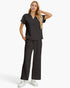 A woman modeling a dark charcoal gray two-piece scrub set consisting of a short-sleeve polo-style top and matching wide-leg pants, paired with white sneakers against a light gray studio background.