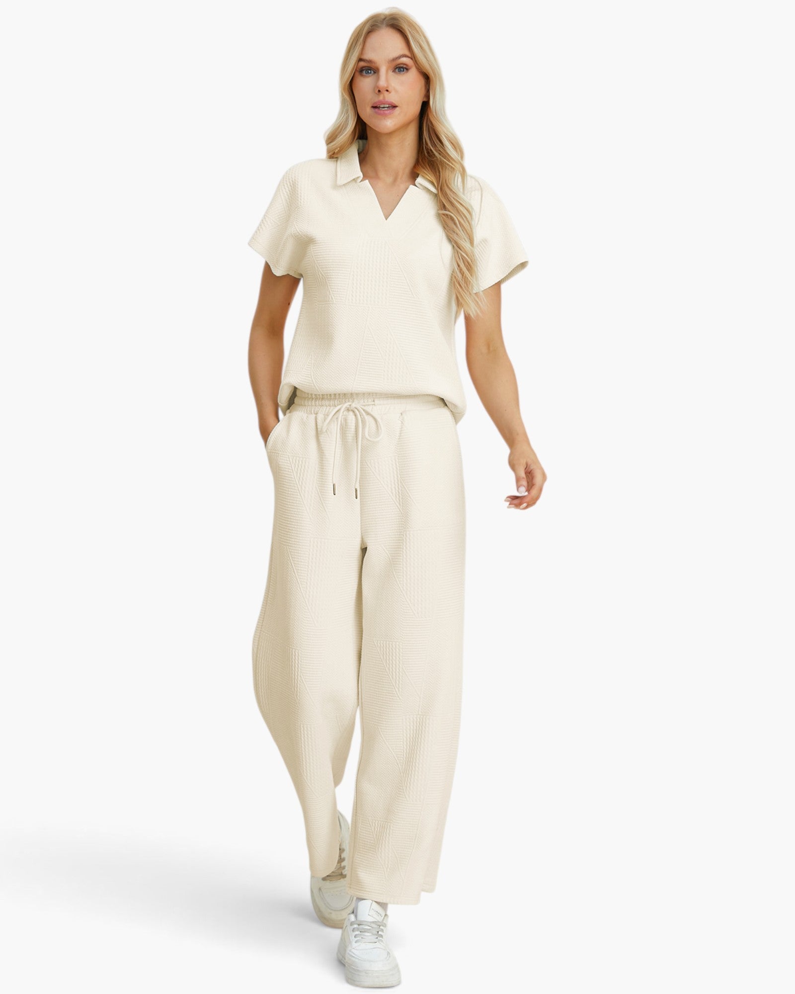 A woman modeling a matching cream-colored two-piece lounge set consisting of a short-sleeve collared top and wide-leg drawstring pants, paired with white sneakers against a plain white studio background.