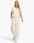 A woman modeling a matching cream-colored two-piece lounge set consisting of a short-sleeve collared top and wide-leg drawstring pants, paired with white sneakers against a plain white studio background.