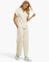 A woman modeling a matching cream-colored lounge set consisting of a short-sleeve polo shirt and wide-leg sweatpants, paired with white sneakers, against a plain white studio background.