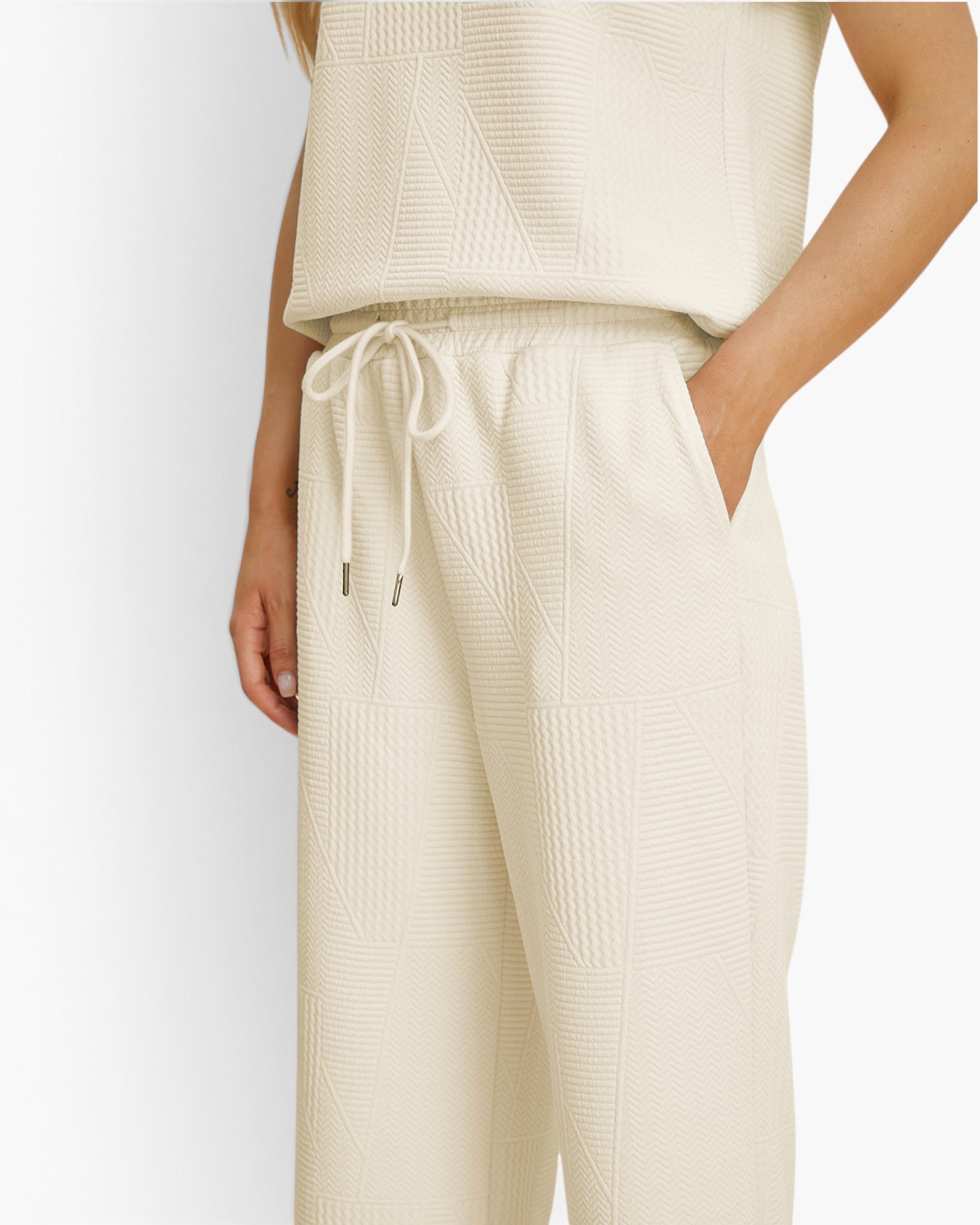 A close-up of a model wearing textured cream-colored knit pants with a drawstring waist and a matching sleeveless top against a plain white background.
