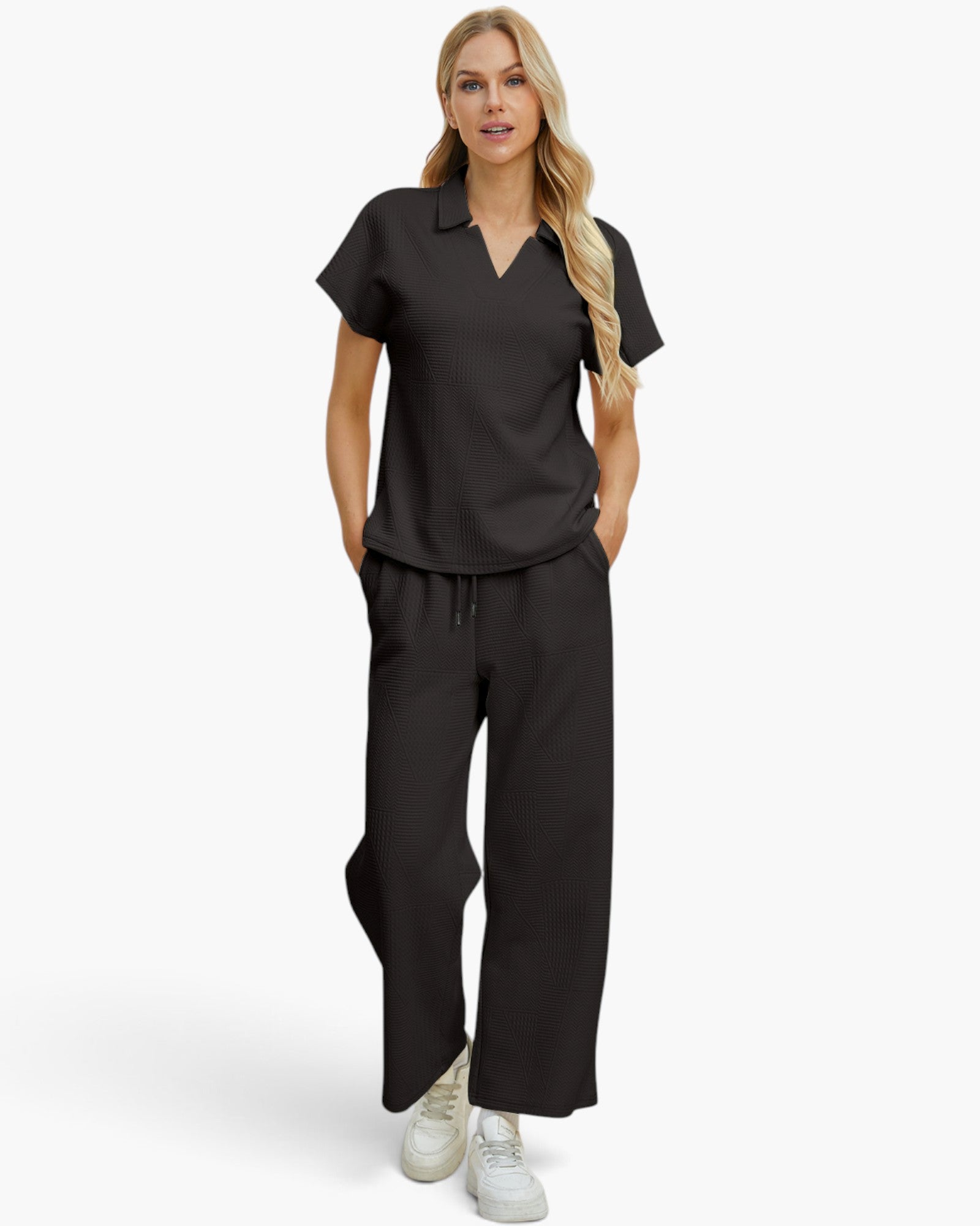 A woman modeling a matching black scrub set consisting of a short-sleeve V-neck top and wide-leg pants, paired with white sneakers, against a light gray studio background.
