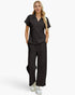 A woman modeling a matching black scrub set consisting of a short-sleeve V-neck top and wide-leg pants, paired with white sneakers, against a light gray studio background.