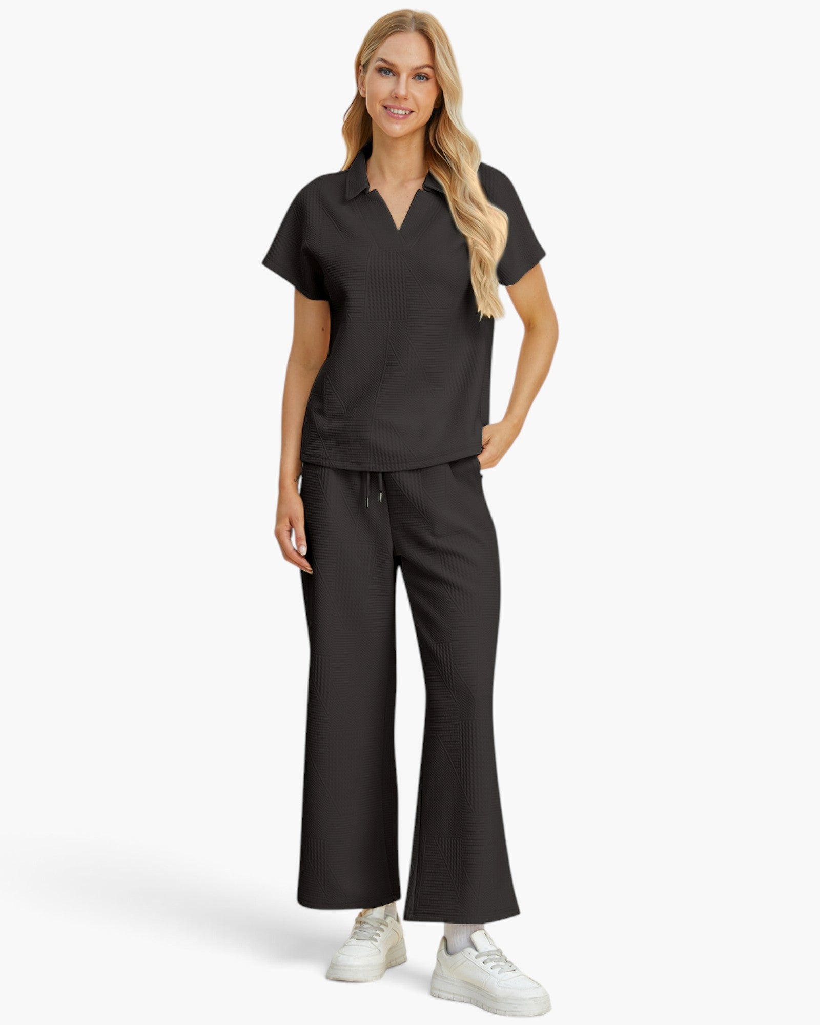 A woman modeling a black short-sleeve scrub top and matching black wide-leg scrub pants with white sneakers, photographed against a clean light gray studio background.