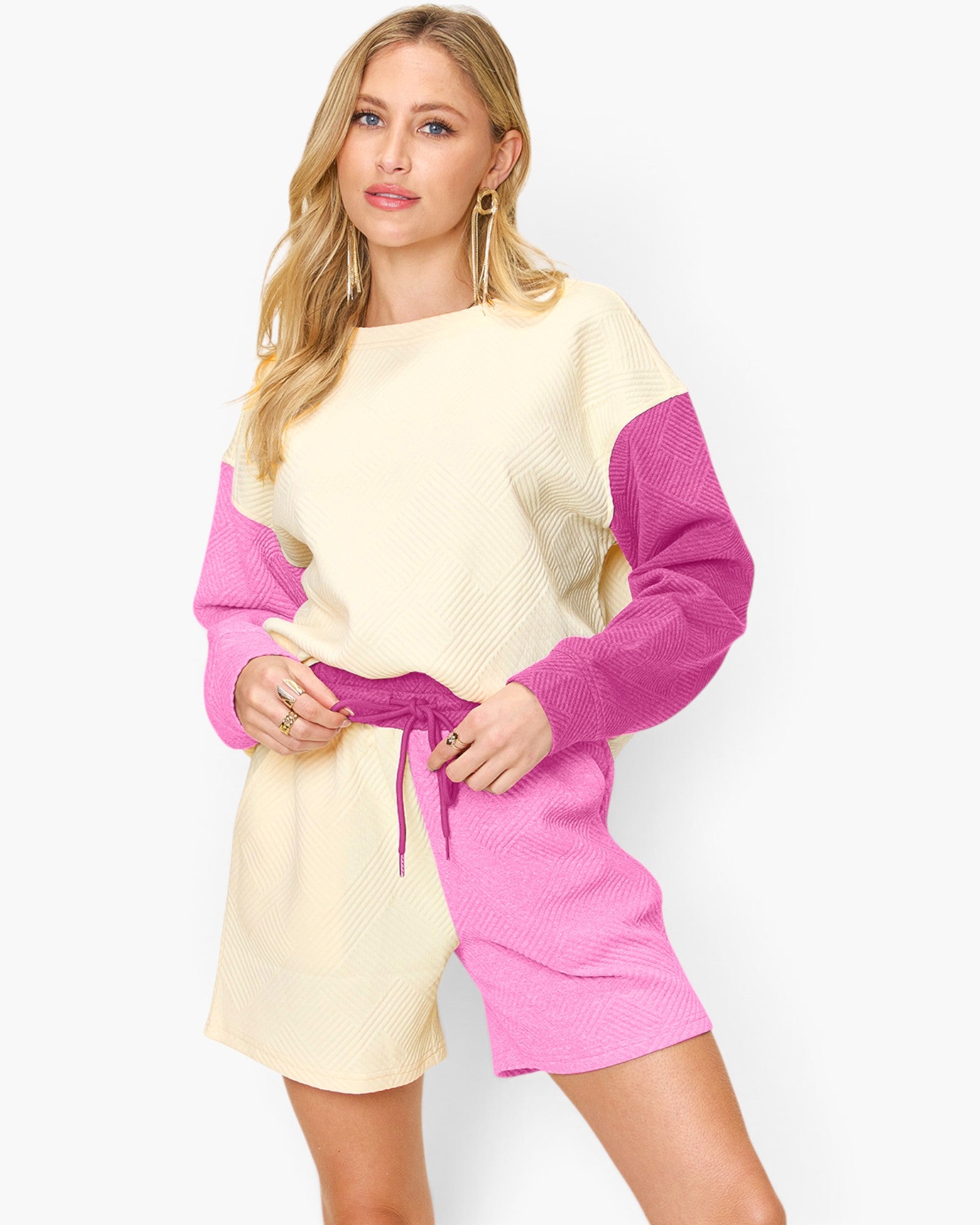 A woman modeling a color-blocked knit romper with a cream body and sleeves, and bright pink shorts and side panels, featuring a matching pink drawstring belt.