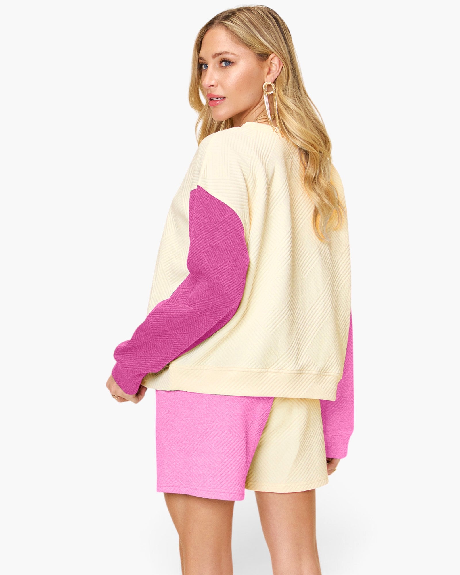 A woman modeling a color-blocked knit sweater and matching shorts set. The set features a cream-colored body with vibrant magenta-pink textured sleeves and shorts, shown from a three-quarters back view against a plain white studio background.