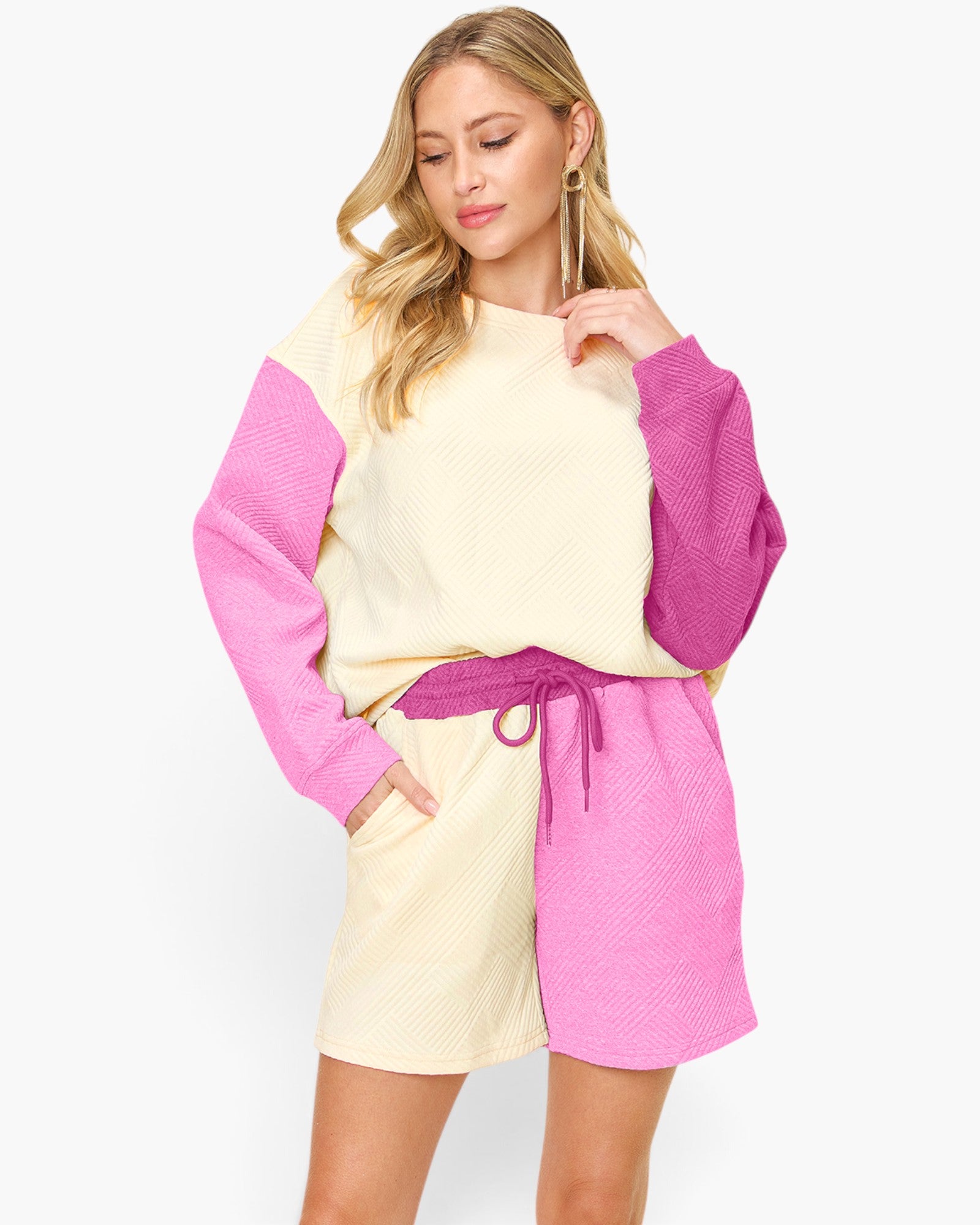 A woman modeling a color-blocked waffle-knit sweatshirt and shorts set. The set features a cream-colored body with bright pink sleeves and a matching pink drawstring belt.