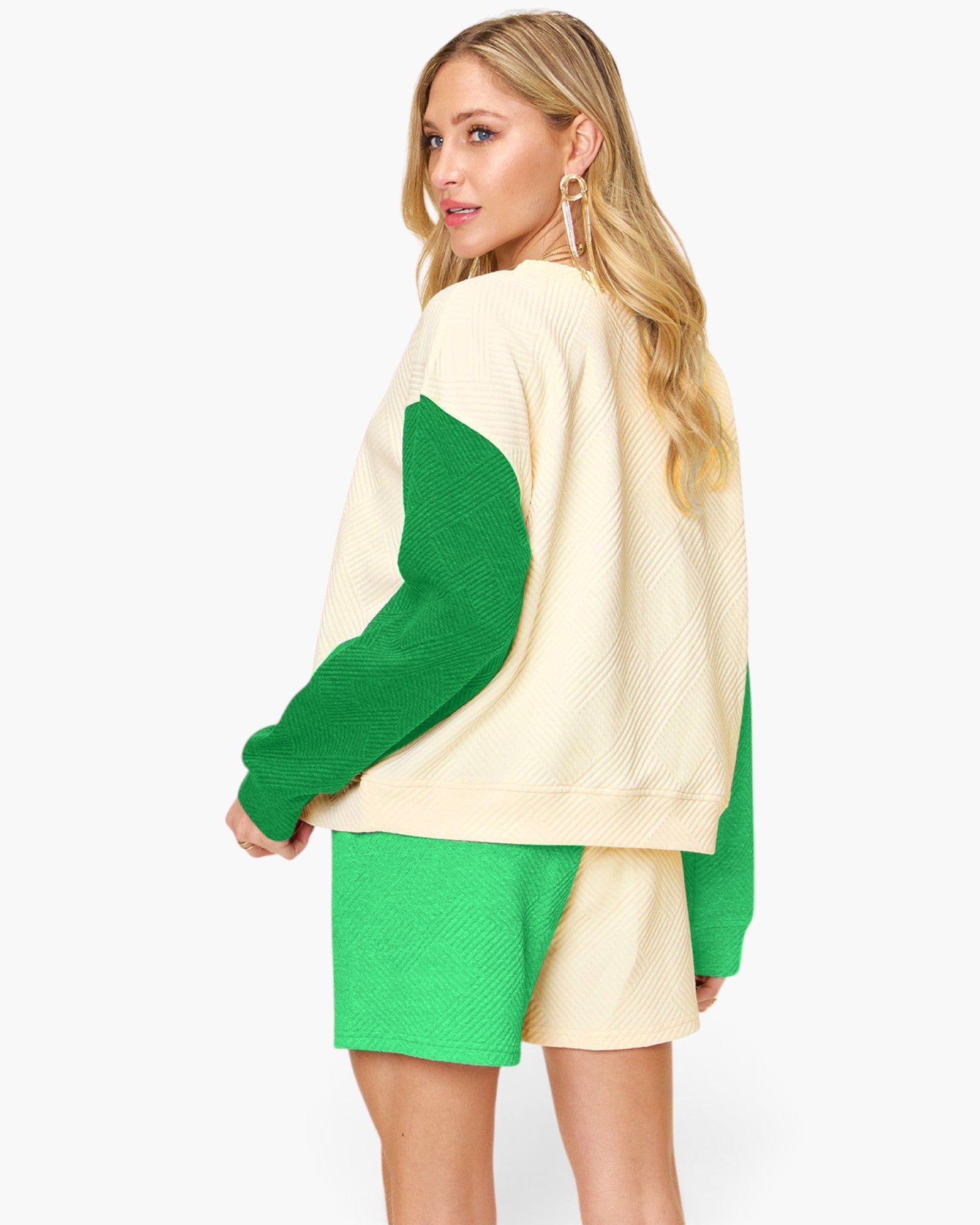 A woman modeling a color-block knit sweater and matching shorts set. The set features a cream-colored body with vibrant green sleeves and side panels, paired with matching green shorts, shot against a plain light gray background.