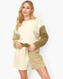 A woman modeling a color-blocked knit jumpsuit with a cream body, olive green sleeves, and matching olive green shorts with a drawstring waist, set against a plain light gray background.