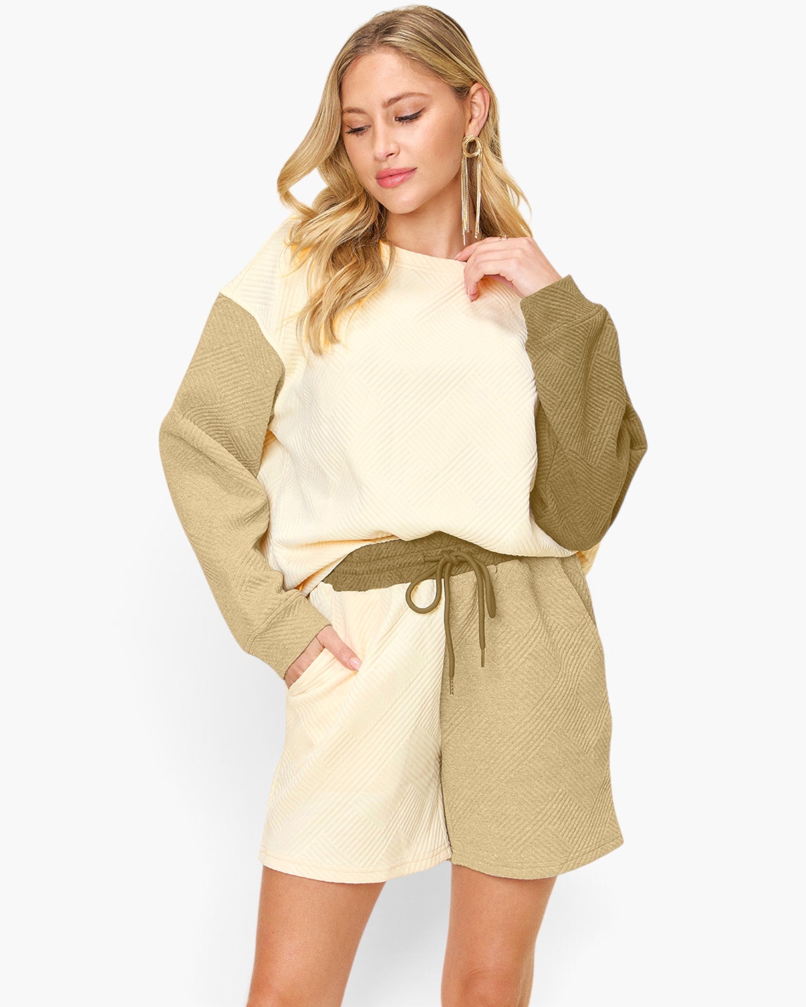 A woman modeling a color-blocked knit jumpsuit with a cream body and olive green sleeves and shorts, featuring a matching olive green drawstring belt, against a plain light gray background.