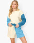 A woman modeling a color-block knit jumpsuit with a cream body and sleeves, and bright blue contrast panels on the sleeves and shorts, featuring a matching blue drawstring belt.