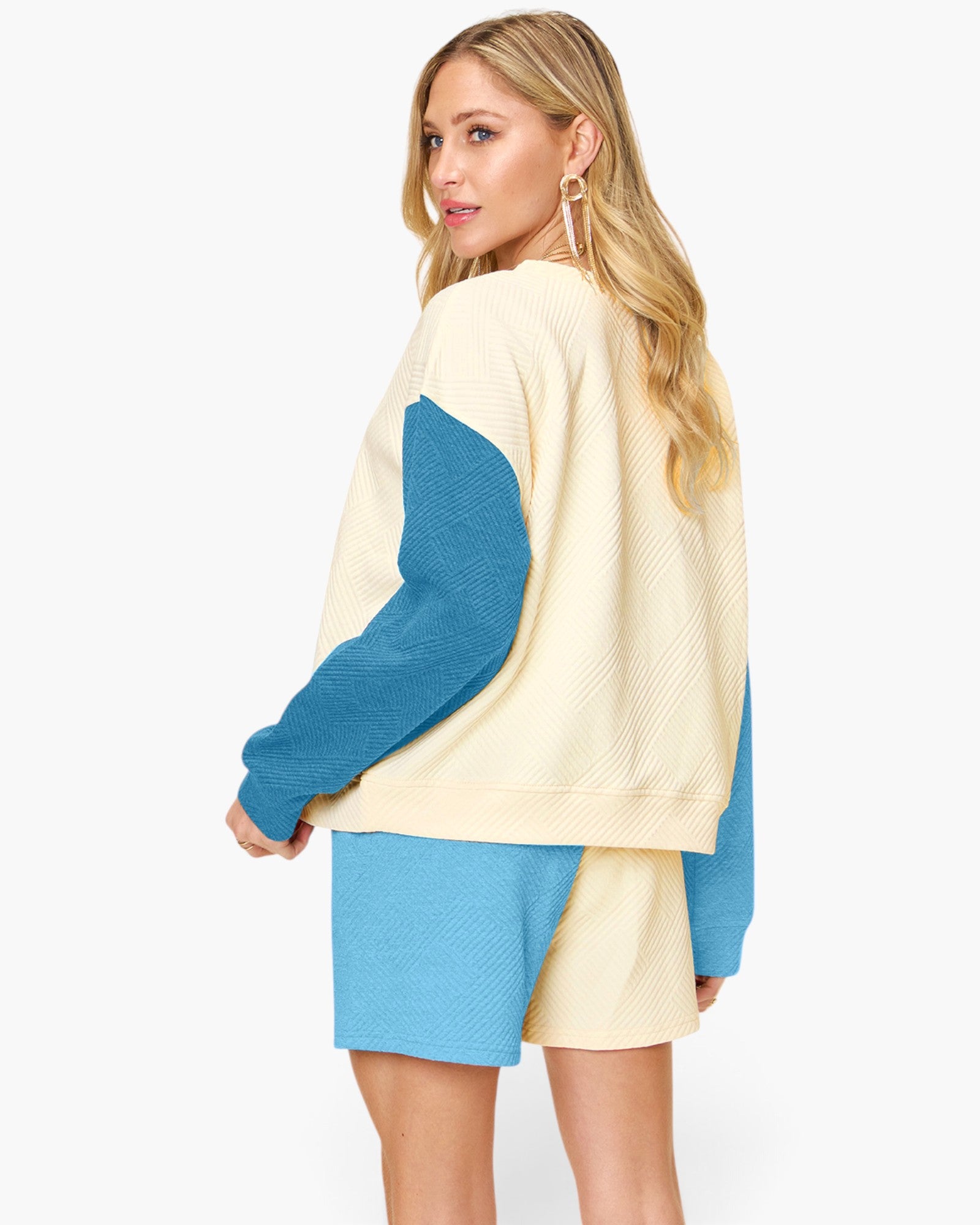 A woman modeling a color-blocked knit sweater and matching shorts set. The set features a cream-colored body with bright blue sleeves and shorts, shown from a three-quarters back view against a plain white studio background.