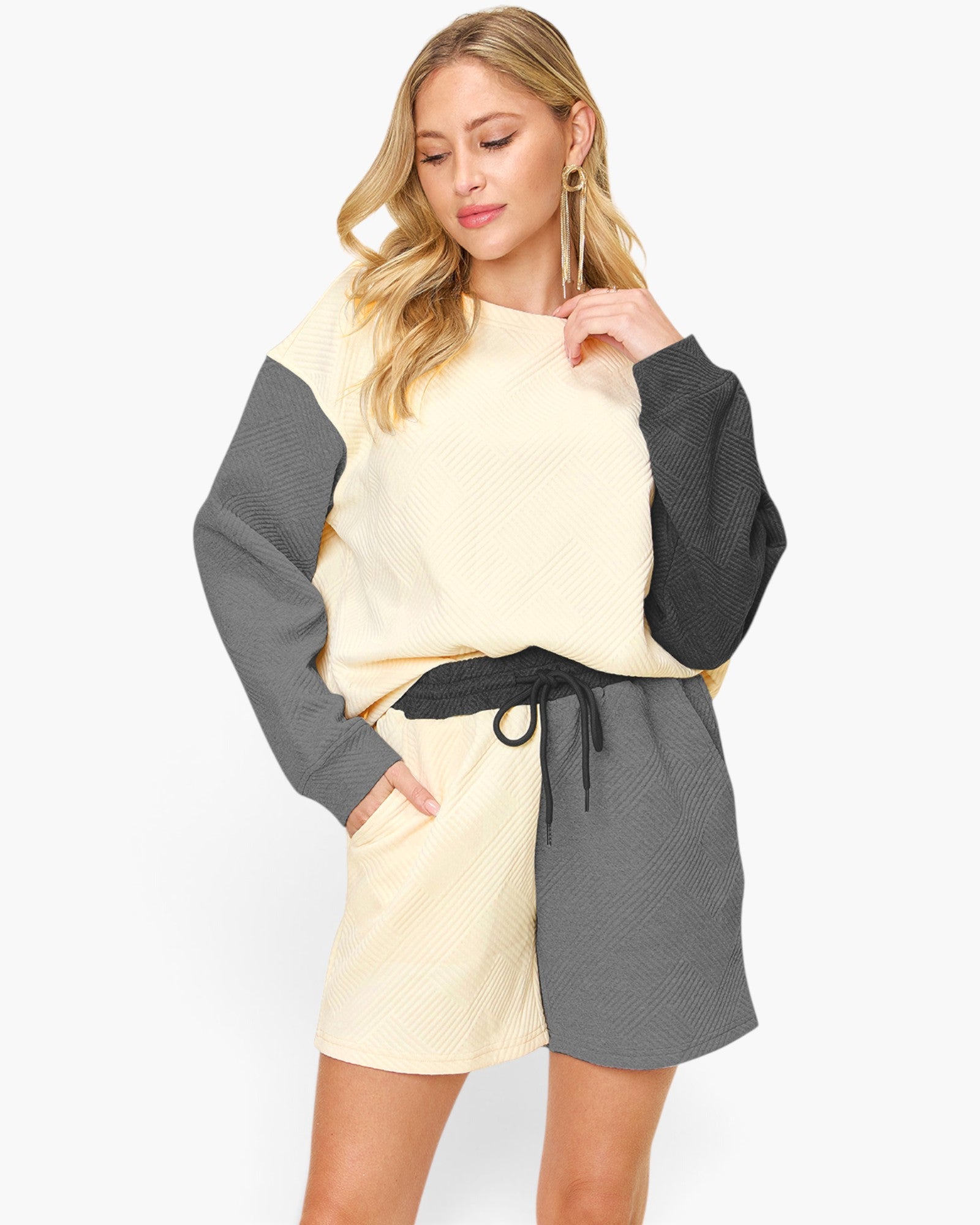 A woman modeling a color-blocked waffle-knit sweatshirt and shorts set. The set features a cream-colored body and shorts with dark gray sleeves and side panels, accented by a black drawstring belt.