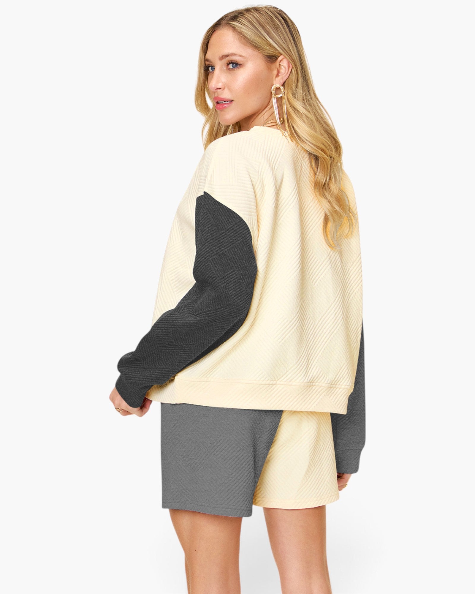 A woman modeling a color-block knit sweater with a cream body and dark charcoal sleeves, paired with matching gray shorts, against a plain white studio background.