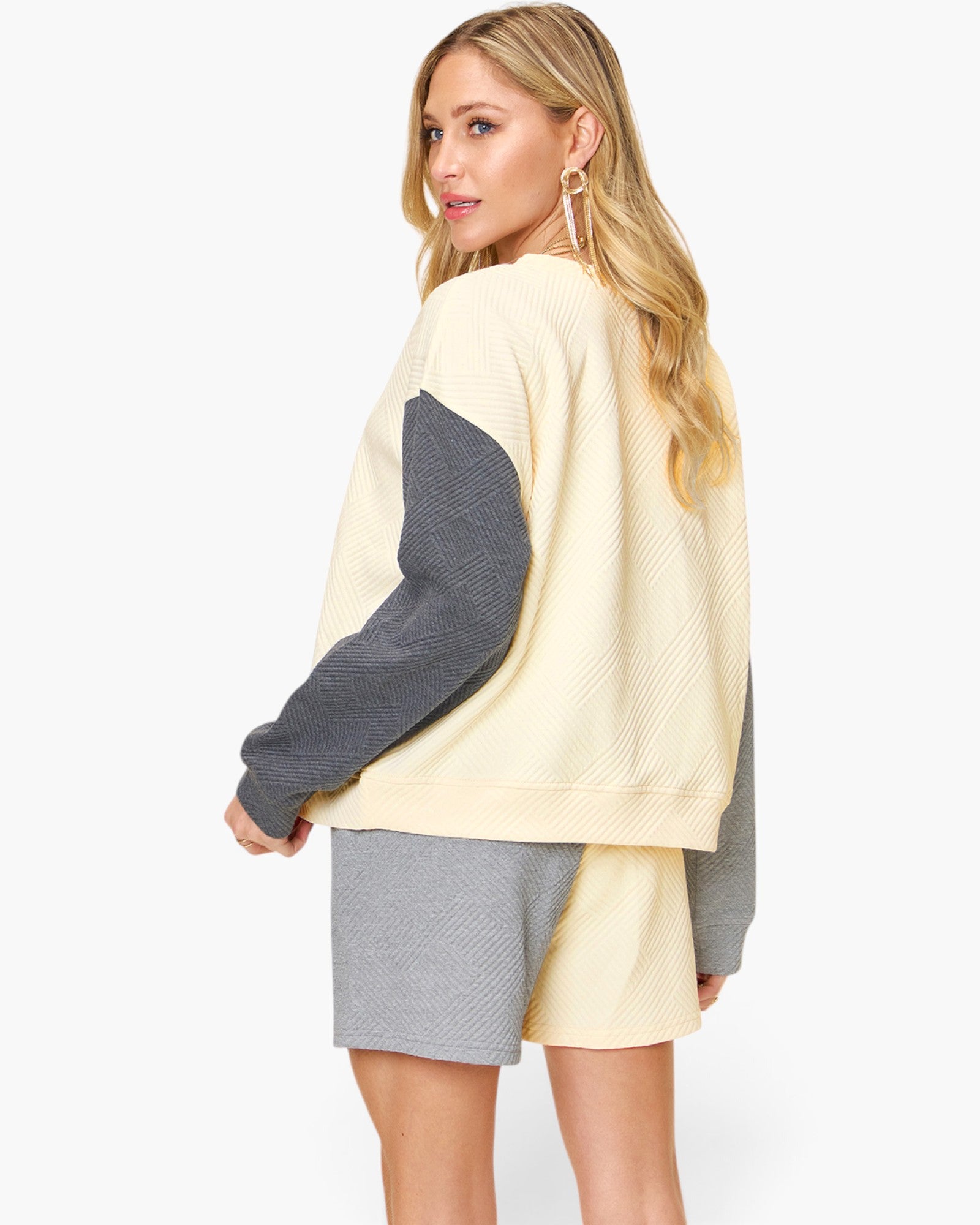 A woman modeling a color-blocked knit sweater with a cream body and dark gray textured sleeves, paired with matching cream and gray shorts, against a plain white background.