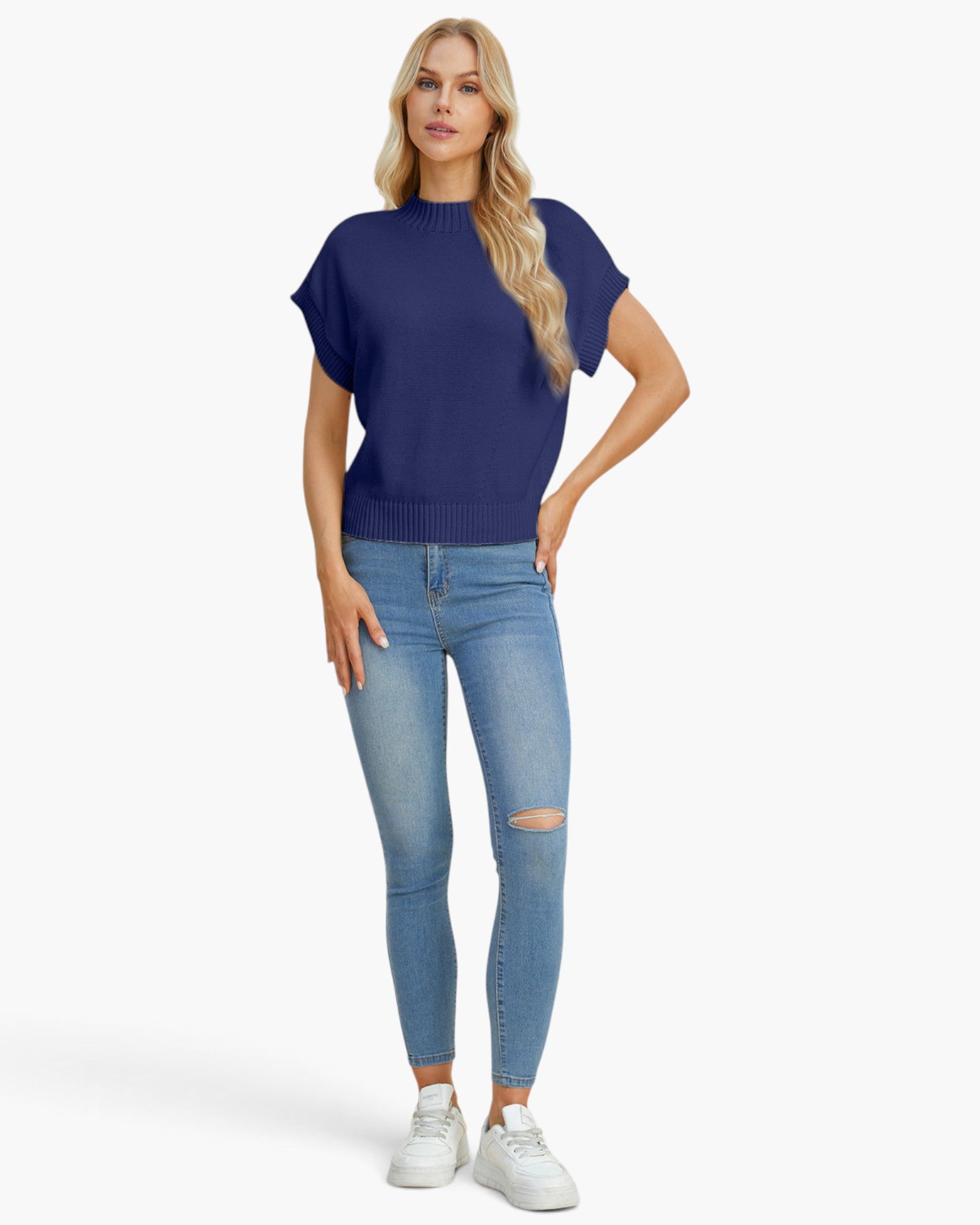 A woman modeling a short-sleeve navy blue knit top with light-wash distressed skinny jeans and white sneakers, standing against a plain light gray studio background.