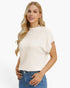 A blonde woman wearing a cream-colored knit sweater with short raglan sleeves and blue jeans, standing against a plain white studio background.