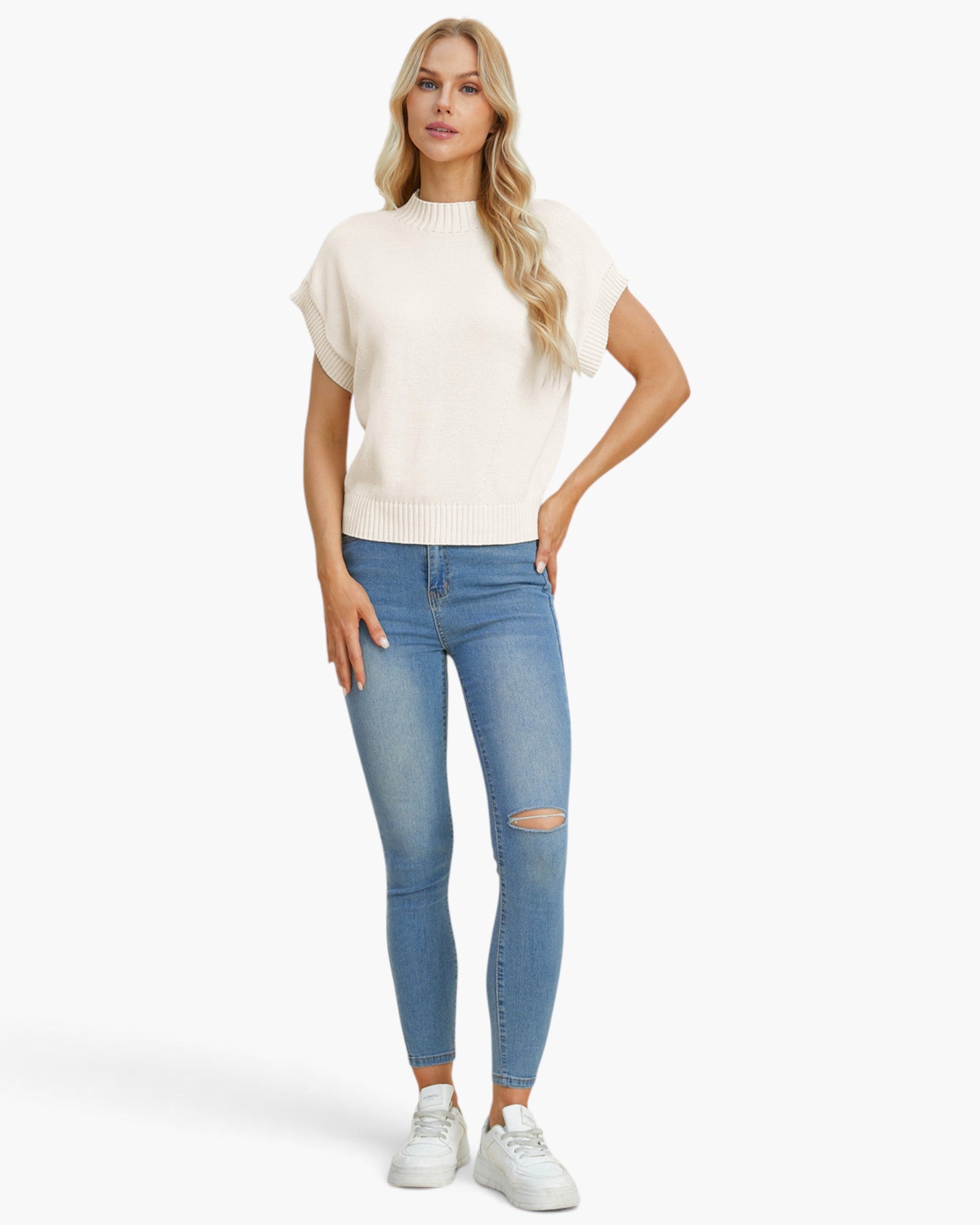 A woman modeling a cream short-sleeve knit sweater and light blue skinny jeans with distressed knees, paired with white sneakers, against a plain white studio background.