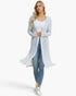 A woman modeling a long, open-front knit cardigan with a textured, marled pattern in white and light blue, worn over a white tank top and blue jeans against a plain white background.